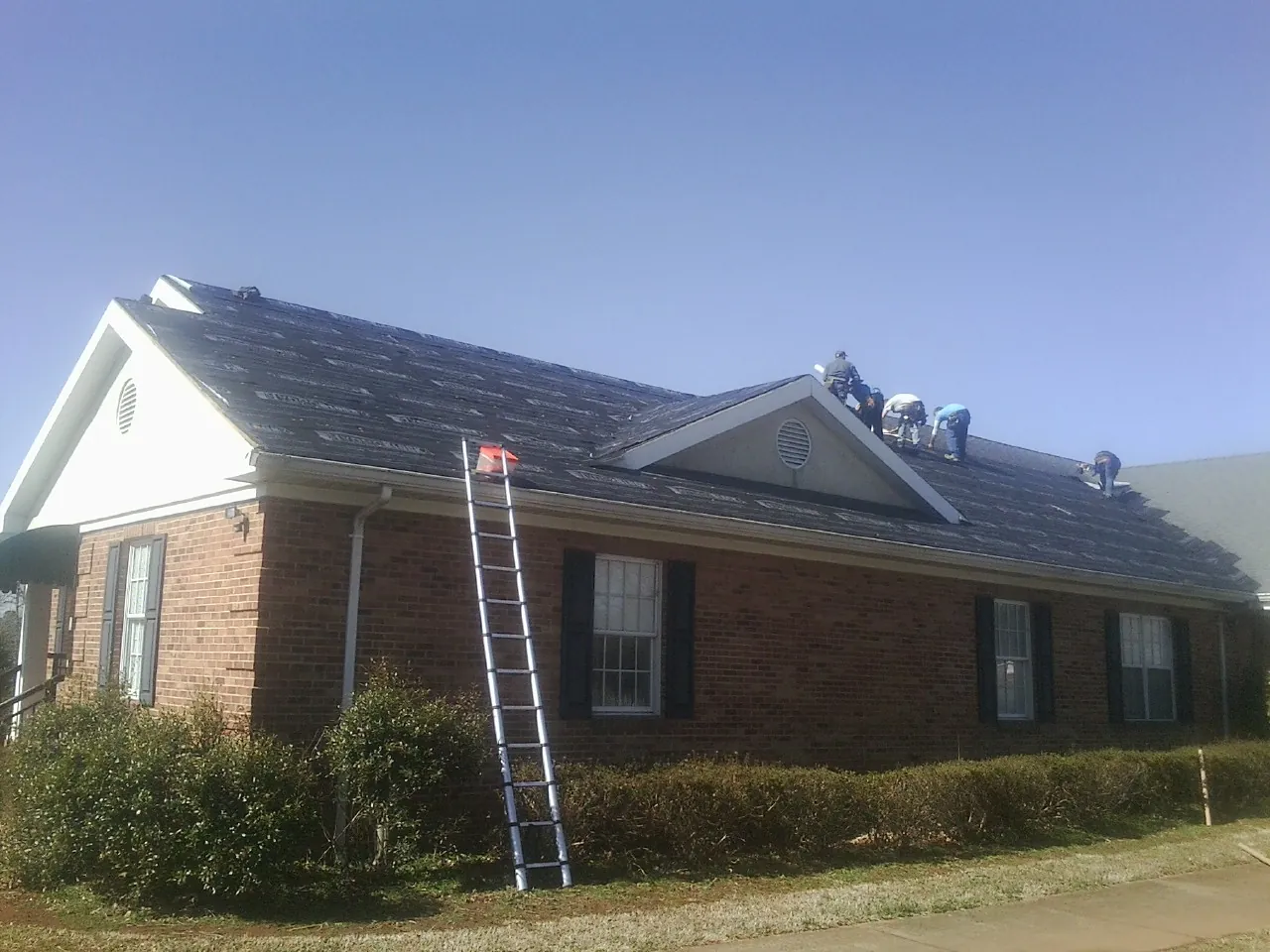Crew tearing shingles off a brick ranch roof with a ladder set — Metal Roof Repair work by our crew in Arvilla
