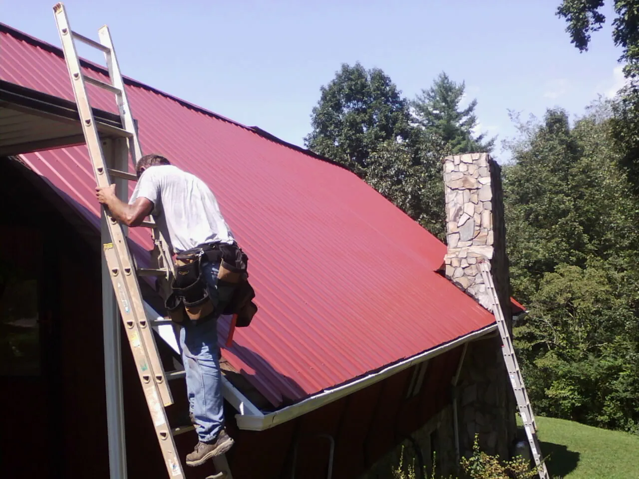 Roofer on a ladder beside a red metal roof and stone chimney — Soffit & Fascia Repair by Roofing Brothers in Arvilla